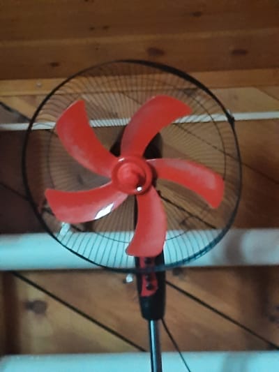 The image shows a close-up, eye-level shot of a red oscillating fan. The fan has black blades and a black protective grill. It is positioned in front of a wooden wall. The fan is the primary subject, and its vibrant red color stands out against the darker background. There is no indication of people or any specific activity taking place, aside from the fan's stationary position. The overall environment appears to be indoors, possibly a room or a workshop. The location of Antonovka, Russia, is not discernible from the visual information provided in the image.