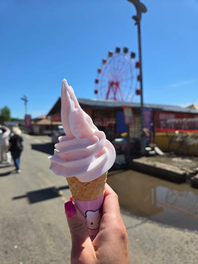 The image, rotated 90 degrees clockwise, prominently features a person's hand holding a pink soft-serve ice cream cone against a clear blue sky. The ice cream is a pale pink hue, swirled elegantly atop a classic waffle cone. A pink paper wrapper, adorned with the iconic "Hello Kitty" character, partially covers the cone. The hand holding the ice cream has fingernails painted with a vibrant fuchsia nail polish.

In the blurred background, a large red Ferris wheel is visible on the left side, indicating an amusement park or carnival setting. Various other indistinct buildings and structures further suggest a recreational area or boardwalk. The ground below appears to be a paved surface, and highly blurred figures of people can be seen in the distance, suggesting other visitors enjoying the outdoor environment.

The scene takes place during bright daylight, evident from the vivid blue sky and clear illumination. The overall impression is one of a sunny day spent at a lively outdoor attraction. The visible text on the wrapper is "Hello Kitty." The location context provided is Jiejinkou, China.