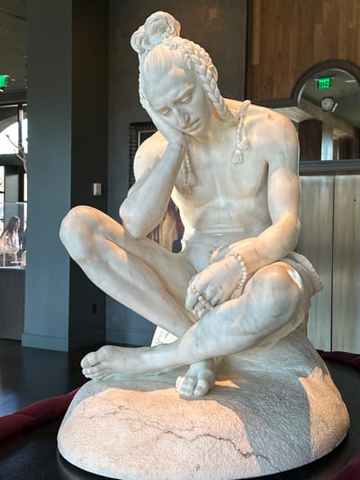 A marble statue depicts a reclining figure with a contemplative pose. The person has their head resting on one hand, with their body partially draped over a rough-hewn stone base. Braided hair adorns the head, and beaded strands are visible on the body. The statue showcases detailed musculature and anatomy, capturing the essence of classical sculpture. The backdrop includes wooden and gray panels, with an illuminated exit sign visible near the top left corner.