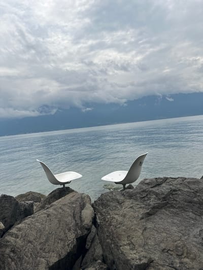 The image depicts a static outdoor scene located on the rocky shore of Lake Geneva in Vevey, Switzerland, under an overcast sky. In the foreground, two modern, white, shell-shaped chairs are individually mounted on large, dark grey boulders. The chairs are positioned facing each other, approximately equidistant from the viewer, appearing to hover slightly above the rock surfaces. The chair on the right exhibits faint, dark, indistinct markings on its upper backrest. The tranquil, blue-grey waters of Lake Geneva extend from the immediate rocky shoreline into the mid-ground. A distant mountain range, partially obscured by low-hanging clouds, defines the horizon. The upper portion of the frame is dominated by a dense, multi-layered cloud cover in shades of grey and white. No individuals are present in the scene.