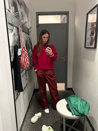 A woman stands in a fitting room, taking a mirror selfie. She is wearing a red quarter-zip sweatshirt and maroon pants. Her hair is brown and long, and she has on glasses. In the foreground, there are two light green clogs on the floor. A round stool with a green jacket draped over it sits to the right. The fitting room has grey walls, a dark grey door, and a floor covered in dark speckled carpet. A rack of clothes hangs on the wall to the left, with a striped bag hanging from a hook. A framed poster is on the wall to the right. This scene is set in a fitting room in the city of Kiryat Ekron, Israel.