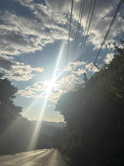 A sun-drenched road stretches ahead, flanked by dense greenery on the right and telephone poles on the left. Power lines crisscross the sky, partially obscured by fluffy white clouds. The sun shines intensely, creating a bright glare and silhouetting distant mountains in a soft, hazy light. The asphalt reflects the sunlight, adding to the overall brightness of the scene.