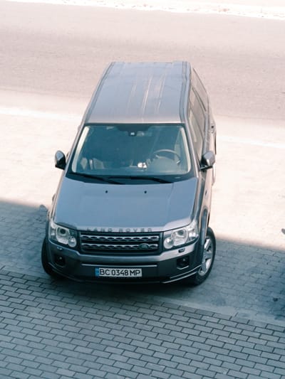 An aerial view captures a dark gray Land Rover Discovery Sport parked on a gray brick-patterned paved surface adjacent to a light-colored asphalt road. The vehicle's license plate, BC 0348 MP, is visible. The car is angled towards the bottom right of the frame. A shadow from an unseen object falls across the front left of the vehicle and the paving stones. The scene is located in Pustomyty, Ukraine.