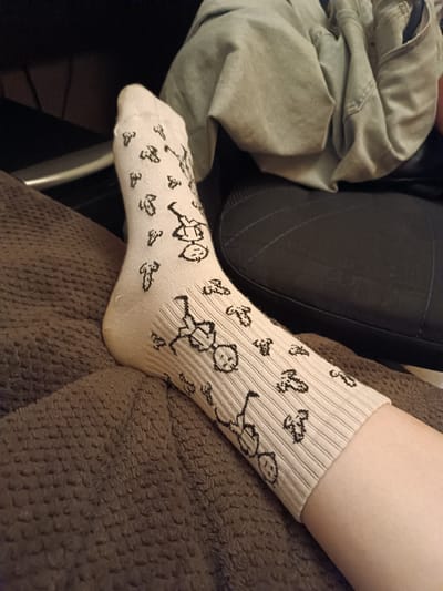 The image shows a close-up of a person's lower leg wearing a light beige sock with a repeating pattern of black stick-figure drawings of what appear to be people. The sock is ribbed and covers the foot and ankle, extending up the calf. The background is blurred and includes a dark surface, possibly a chair, and a crumpled piece of light-colored fabric. The person is resting their leg on a dark brown textured blanket. There is no indication of the location within the city of Kozmodemyansk, Russia.