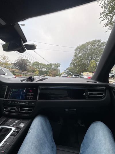 The image presents a first-person perspective from the passenger seat of a dark-interior vehicle, identified as a Porsche Macan, looking forward onto a residential street in Newton, United States. The sky is overcast.
Inside the vehicle, the black dashboard features a central infotainment screen displaying a navigation map on its left side and a radio interface on the right, tuned to "107.9 FM HD1" playing "Students of the Dream Nation." A circular gauge is mounted on the dashboard's upper center. The center console contains numerous buttons and a gear selector. The lower part of the image shows the legs of an occupant, clad in light blue ripped jeans, with dark shoes partially visible on the black floor mat.
Outside the vehicle, a line of traffic is stopped on the street. A yellow school bus, displaying "SCHOOL BUS" lettering, is prominently positioned on the left side of the road. Overhead, multiple power lines traverse the sky. Abundant trees with green foliage line both sides of the street, partially obscuring residential structures. A dark fence is visible running along the left side of the road.