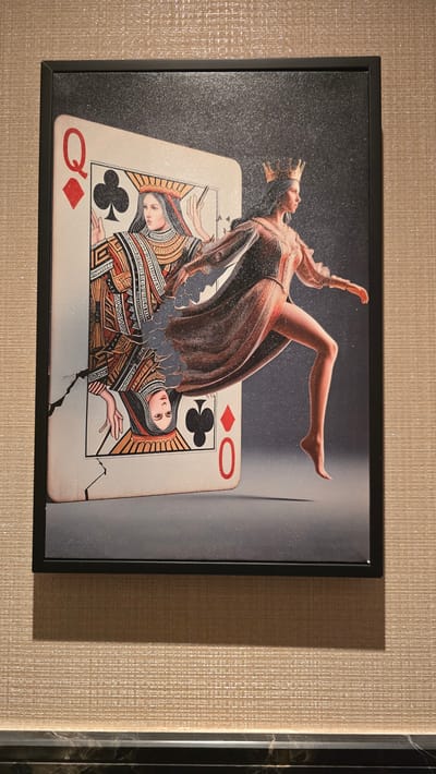 A framed painting is displayed on a textured beige wall. The painting depicts a surreal scene where a female figure, dressed in a sparkling, peach-colored gown and wearing a golden crown, is emerging from a giant playing card, specifically the Queen of Spades. The playing card is partially torn, revealing another representation of a queen on the other side. The background of the painting is dark and speckled, giving the impression of a starry night. The wall it hangs on has a woven texture and is illuminated by a light source from above, casting a shadow of the frame onto the wall below. The painting is bordered by a thin, black frame.
