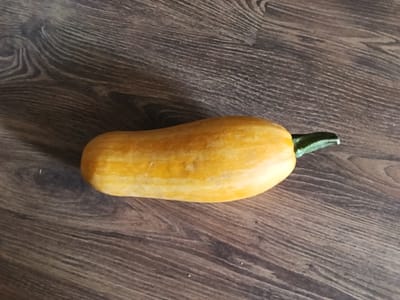 A single, elongated, yellow squash lies horizontally on a dark brown, wood-grained surface. The squash is smooth with subtle lighter yellow striations running lengthwise. One end of the squash tapers to a short, dark green stem. The surface it rests on has a visible wood grain pattern with variations in tone and texture. There are no people or other objects present in the image. The scene is static and appears to be an indoor still life. There is no information within the image to suggest a location in Drahichyn, Belarus.