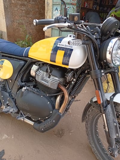 The image shows a Royal Enfield motorcycle parked on a dirt surface in Shella ward, Kenya. The motorcycle, oriented from a front-left three-quarter view, features a yellow and white fuel tank with a dark blue central stripe and 'ROYAL ENFIELD' branding. A circular yellow side panel on the side displays '650' in black. It has a dark blue seat, exposed air-cooled engine cylinders, a black engine case, and a gold-toned exhaust pipe with a black perforated heat shield. The front wheel is a wire-spoke type with a knobby tire, and the front fender is white. A round chrome-bezeled headlight and clear LED turn signals are visible. In the background, to the left, is an unpainted wall. To the right, a building with a faded blue wooden door or shutter is partially seen. Inside this structure, a person in a dark shirt is bent over, largely obscured by the motorcycle's handlebars, engaging in an activity within what appears to be a shop. Shelves containing various items are visible behind the person. The scene is illuminated by natural daylight.