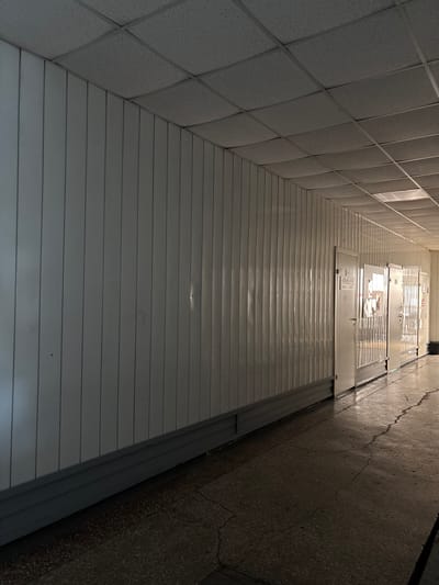 A photograph, taken from a low angle, shows a long, empty hallway with white paneled walls and a drop ceiling. The hallway extends into the distance, with several doors visible on the right side. The floor is made of dark, cracked concrete. The lighting in the hallway is dim, with a few fluorescent lights visible in the ceiling. The overall impression is one of a sterile and utilitarian environment.