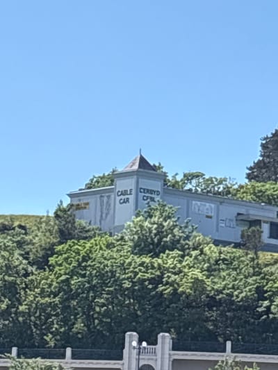 The image depicts the Llandudno Cable Car station, partially obscured by dense green foliage, under a clear blue sky. The station is a multi-story structure with a pointed roof, bearing the words "CABLE CAR" and "CERBYD CFBL" vertically on its facade. A portion of a decorative balustrade or railing is visible on the right edge of the frame. The environment appears to be a hillside location within Llandudno, United Kingdom, due to the station's location and signage in English and Welsh.