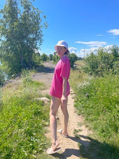 A young woman with blonde hair, wearing a pink t-shirt and a white cap, stands on a path in a grassy area with trees. She is facing towards the viewer, with her head tilted slightly. The sun is shining brightly, casting shadows on the ground, suggesting it is daytime. The sky is a clear blue with a few white clouds. In the background, there are more trees and a rocky, uneven landscape. A cell tower can be seen in the distance. The woman appears to be relaxed and enjoying the outdoors. The image is taken in Belozersk, Russia.