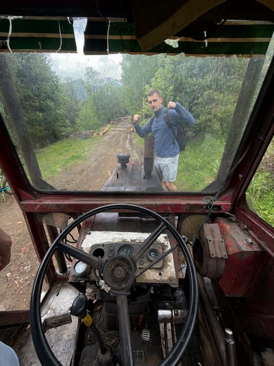 The image shows the interior of an old, red vehicle, possibly a truck or tractor, with a steering wheel and dashboard visible. The dashboard is worn and covered in dirt. Through the cracked and dirty windshield, a reflection of a man in a blue shirt and light-colored shorts is visible. The background seen through the windshield and reflected is a forest. The location is Рилски манастир, Bulgaria. The interior of the vehicle appears derelict and unused. The man is standing outside the vehicle, seemingly posing for the photo.