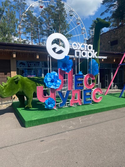 An outdoor scene in Mistolovo, Russia, captures a bright, sunny day with a Ferris wheel visible in the background. In the foreground, a large, green, artificial rhinoceros stands on a green platform. Behind it, a colorful three-dimensional sign spells out "ЧУДЕС" (Chudes) with large, block letters in pink and blue. The letters are decorated with large, blue paper flowers. To the right of the sign, a tall, green, artificial giraffe sculpture extends upwards. The building behind the sign has a wooden facade and large windows. The ground is paved with asphalt.