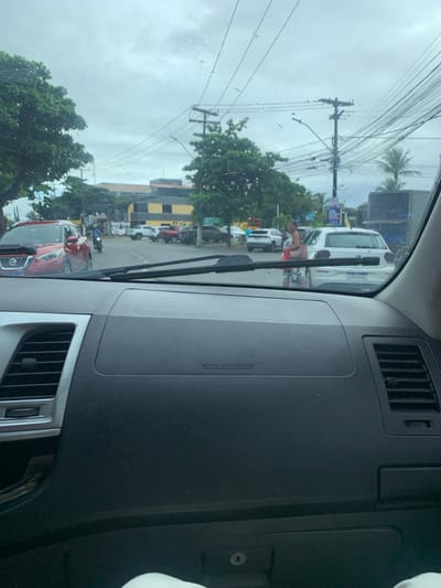 The image displays an urban street scene in Lauro de Freitas, Brazil, observed from the interior of a stationary vehicle. The foreground shows the vehicle's dark grey dashboard, complete with an "SRS AIRBAG" emblem and air vents on both sides. Windshield wipers are positioned horizontally across the base of the windshield, which has visible water spots and reflections.
The sky is overcast. On the asphalt roadway, a red Nissan Kicks SUV is partially visible on the left, followed by a motorcyclist wearing a blue top and black helmet. To the right, a white Volkswagen Polo hatchback, displaying Brazilian Mercosul license plate YAC5A62, is present. An adult female pedestrian is seen in the mid-right, captured mid-stride, wearing a patterned dress, carrying a light-brown shoulder bag and a red item in her left hand, positioned near the rear of the white hatchback.
Multiple other vehicles, including white and red pickup trucks and dark-colored SUVs, are parked or stationary in the mid-ground. A dark truck is partially visible on the far right. The environment features green trees lining the street and a distinctive two-story building in the background with a yellow and dark-grey facade. Numerous utility poles supporting a dense network of overhead power lines and communication cables dominate the upper portion of the frame.