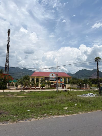 The image depicts a street-level view of a building with a red roof, likely a government or administrative building, in Dien Tho, Vietnam. The building is yellow with white trim and has a flagpole in front. Several flags are visible on poles in front of the building. The building is surrounded by a fence and trees. To the left of the building, a tall, lattice-style telecommunications tower is visible, along with some red-flowering trees. In the background, there are green hills. The foreground shows a grassy area with a paved road in front. The sky is partly cloudy with large, white cumulus clouds. There are overhead power lines crisscrossing the scene. The overall impression is of a rural or semi-urban setting.
