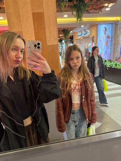 A selfie is taken in front of a mirror, reflecting three people. The foreground shows a woman holding an iPhone with a clear case decorated with bows. She wears a black jacket with white stripes and has blonde hair. The reflection shows a younger girl with blonde hair, wearing a brown jacket, a pink top, and blue jeans. Further back in the reflection is a person wearing a leather jacket and blue jeans. The location appears to be a shopping mall in Ol'ginskaya, Russia, based on signage and architectural style in the background. The environment includes storefronts, displays, and greenery. The interaction is a group taking a picture of themselves in a mirror at a shopping center.