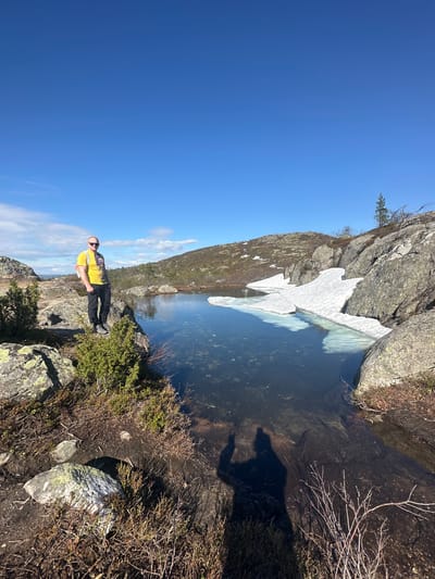 A man wearing a yellow shirt and black pants stands on a rocky ledge overlooking a small body of water. Patches of ice float in the water near a rock face. The sky is clear blue. The location is likely near Kandalaksha, Russia, given the landscape and presence of glacial features like the ice patch. The man is facing the camera and appears to be posing. The environment consists of rocks, low-lying vegetation, and a small lake or pond. The sun is shining brightly, casting a shadow of the man onto the ground near the water's edge.
