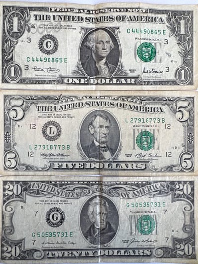 This media file contains a close-up, overhead view of three United States dollar bills placed side-by-side. From left to right, they are a one-dollar bill, a five-dollar bill, and a twenty-dollar bill. The bills are slightly creased and worn, indicating they are in circulation.
The one-dollar bill features a portrait of George Washington. Visible text includes "FEDERAL RESERVE NOTE," "THE UNITED STATES OF AMERICA," "ONE DOLLAR," a serial number "C44490865 E," and signatures of the Treasurer and Secretary of the Treasury.
The five-dollar bill displays a portrait of Abraham Lincoln. Visible text includes "FEDERAL RESERVE NOTE," "THE UNITED STATES OF AMERICA," "FIVE DOLLARS," a serial number "L27918773 B," and signatures.
The twenty-dollar bill shows a portrait of Andrew Jackson. Visible text includes "FEDERAL RESERVE NOTE," "THE UNITED STATES OF AMERICA," "TWENTY DOLLARS," a serial number "G50535731 E," and signatures.
The bills are laid out horizontally on a plain, light-colored surface, likely a table or countertop. There are no other objects or environmental details visible in the frame, suggesting a controlled studio or workspace setting. The lighting appears to be bright and even, illuminating the details of the currency clearly. The focus is entirely on the monetary items.