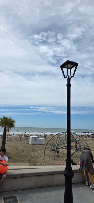 The image shows a view of the beach in Benicàssim, Spain. A black lamppost is prominently positioned in the foreground, stretching horizontally across the frame.
The beach is visible in the background, showing a sandy area with people, beach umbrellas, and a playground structure with children. The ocean is visible in the distance. A palm tree is partially visible near the bottom of the image.
A person wearing a white shirt and orange shorts is seen lying down on a paved area near the bottom right of the image. Other people are present on the sidewalk to the right of the lamppost.
The sky is filled with clouds.