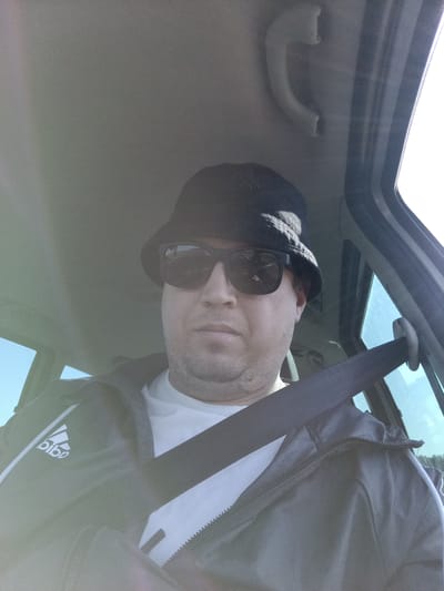 The image shows a man wearing a black bucket hat and sunglasses, seated inside a vehicle. He is wearing a white t-shirt and a black jacket with white accents and the Adidas logo. A black seatbelt is fastened across his chest. The interior of the vehicle, including a grab handle on the ceiling and part of the window frame, is visible. The lighting suggests it is daytime. The exact location within Čistá, Czechia is not identifiable from the image.