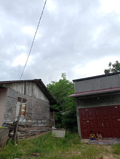 Two young children are sitting on a mattress in front of a dark red double door in Meunasahteungkudigadong, Indonesia. The door is part of a gray building with a dark red roof overhang. To the left is an older, weathered wooden building with a woven exterior. Green vegetation and trees surround the buildings. An electrical wire stretches across the sky, which is overcast with gray clouds. Debris and overgrown grass fill the foreground.