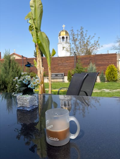 The image shows an outdoor scene, likely a backyard or patio, in Karadjovo, Bulgaria. A black table is the focal point. On the table, there is a glass coffee mug, approximately half-filled with a brown liquid, and two square glass vases. One vase contains small white pebbles and the other contains an arrangement of artificial white and blue flowers. A decorative butterfly is attached to the floral arrangement. In the background, there are two black patio chairs, a wooden fence, and various trees and bushes. A white church steeple with a gold dome is visible in the distance, indicating the location within Karadjovo. The sky is blue with some scattered clouds. The scene is well-lit, suggesting it is daytime. The table surface reflects the objects placed on it, along with the surrounding environment.