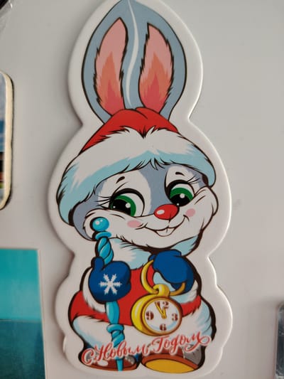 The image features a close-up of a festive, cartoon-style rabbit character, likely a magnet or sticker, set against a plain white background. The rabbit is designed with predominantly white and light blue fur, sporting large, expressive green eyes with long eyelashes, a small red nose, and rosy pink cheeks. Its inner ears are also pink.
The character is adorned in winter holiday attire, including a red and white Santa hat. It wears blue mittens, one of which has a white snowflake emblem. In its left mittened paw, the rabbit holds a light blue staff or scepter with a spiraled design and a spherical top. In its right mittened paw, it clutches a golden pocket watch with a white face and black numerals, indicating the time is set just past midnight. The rabbit has a wide, happy smile, suggesting a joyful demeanor.
Below the rabbit, in decorative red script, the text "С Новым Годом" is visible, which translates from Russian as "Happy New Year." The overall scene portrays a celebratory atmosphere, likely associated with the New Year holiday, given the symbols of the Santa hat, snowflake, and the "Happy New Year" greeting. The location context for this media file is Сусады-Эбалак, Russia.