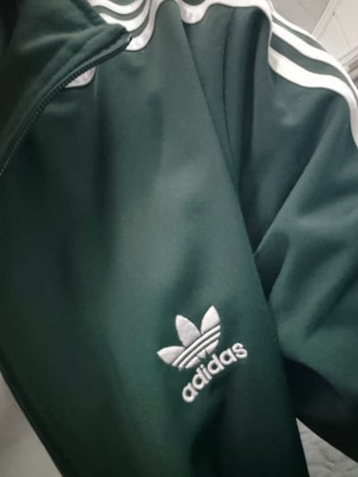 The image shows a close-up view of a dark green Adidas tracksuit jacket. The iconic three white stripes are visible on the sleeve, and the Adidas trefoil logo with the word "adidas" underneath is embroidered in white on the lower part of the sleeve. The jacket appears to be made of a soft fabric, possibly cotton or polyester. The lighting is dim, casting shadows on the fabric, which suggests it might be indoors. There is no information in the image to place it specifically in Lillehammer, Norway.