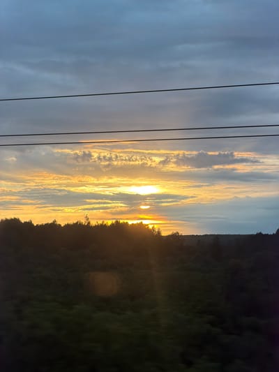 The image shows a landscape at sunset, likely in Arsk, Russia, though no specific identifiable landmarks are visible. A line of power lines runs vertically along the left side of the frame. The sky is filled with a mix of clouds, with the sun setting, casting a golden glow on the clouds. To the right, a dark, silhouetted area suggests the presence of a forest or hillside.
