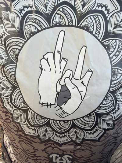 A close-up photograph displays a patterned surface, likely fabric or a flexible material, featuring a large, intricate black and white mandala design. Centrally, a light grey circular area contains an illustration of two stylized hands. The left hand, rendered in white with black outlines, shows the middle finger extended upwards, while the index finger is slightly raised and the remaining fingers are curled. The right hand, also white with black outlines, forms a "V" shape with the extended index and middle fingers, while the thumb and remaining fingers are curled. A dark grey shadow is visible beneath the palm area of the right hand. The right hand appears positioned slightly in front of the left hand. The mandala pattern, visible around the central circle, consists of repeating floral and geometric motifs. At the very bottom of the image, partially visible, is the stylized white text "THC." The image depicts a detail of an object located in El Prat De Llobregat, Spain.