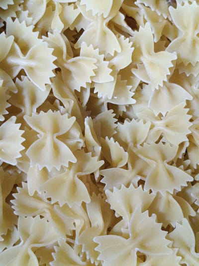 The image is a close-up, overhead shot of a pile of uncooked farfalle pasta, commonly known as bow-tie pasta. The pasta is a pale yellow color and has a slightly textured surface. The pieces are overlapping and arranged randomly, creating a dense, textured pattern. The lighting is even, casting soft shadows that highlight the shapes of the pasta. There are no people or any other objects visible in the image. The background appears to be a neutral surface that blends with the color of the pasta. The image does not contain any information that would indicate a specific location in Martuni, Armenia, or any other place.