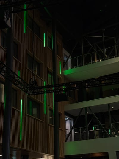 The image features an upward view of a modern building interior. The walls are clad in horizontal wood panels with neon green lights accentuating the windowsills. Overhead, structural beams and scaffolding create a geometric pattern against a dark background. A black, metal stage light tower stands prominently in the foreground, partially obscuring the building's façade. The lighting gives the scene a contemporary, theatrical feel.