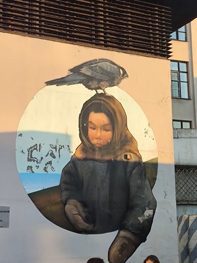 A mural on a building depicts a young person wearing a hooded jacket and mittens, with a bird perched on their head. The person's face is rendered in a realistic style, with a somber expression. The mural is set against a pale background with a hint of blue and a brown circular frame. The image is captured in the warm, golden light of sunset, casting long shadows and highlighting the texture of the wall.

To the right of the mural, a building with multiple windows and a concrete facade is visible. Above the mural, a dark, slatted overhang or structure extends across the top of the wall. The weather appears clear, with the sunset suggesting a calm evening.

On the left side of the mural, a small sign with text is affixed to the wall. The visible text includes "ЕСКОЕ" and "АНА," which are partial words, possibly indicating a location or name. The overall scene has an artistic and perhaps poignant feel, with the child and bird juxtaposed against the urban backdrop. The peeling paint on the wall adds a sense of age and texture to the environment.