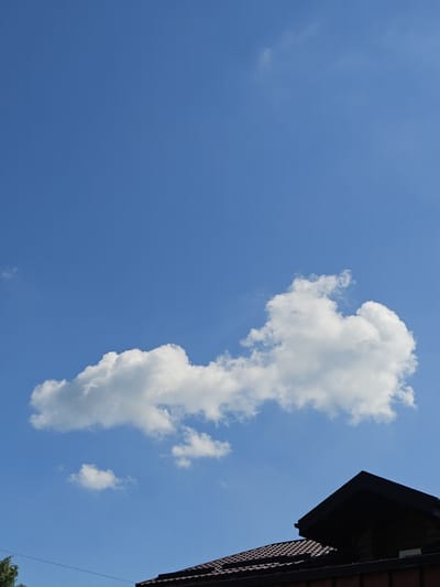 The image depicts a clear blue sky with scattered white cumulus clouds. In the lower right corner, the dark, angular roofline of a building is visible, suggesting a residential structure. A portion of a green tree is present in the lower left corner. The overall scene appears to be an outdoor view, likely from a residential area in Shkurupiivka, Ukraine. There are no people or animals visible in the frame. The image captures a tranquil, daytime atmosphere with a focus on the sky and the upper portion of a building.