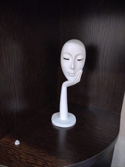 This media file shows a white decorative bust of a woman's face resting on a dark wooden shelf. The bust has closed eyes and its left hand is raised to its cheek, giving it a thoughtful or serene expression. The bust is standing on a white circular base. The shelf is dark brown with a wood grain pattern and appears to be part of a cabinet or built-in shelving unit. There is a small, clear plastic object on the shelf to the left of the bust, possibly a shelf support. The background is a darker, vertically oriented wood grain pattern, suggesting the inside of a cupboard or the back of a shelf. The lighting suggests an indoor setting, possibly during the day, with the light source coming from the front and slightly to the right. There is no visible text in the image. The setting is likely a home or an office in Нижняя Мактама, Russia.