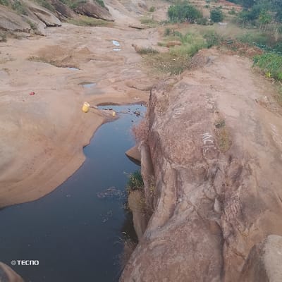 A landscape featuring a rocky terrain with water pooling in depressions. A large rock formation dominates the right side of the image. A yellow container sits near a pool of water. Vegetation is scattered throughout, with denser patches in the background. The area appears dry and arid, with signs of erosion. "TECNO" is visible in the lower left corner.
