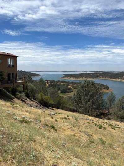 The image depicts a scenic view of Folsom Lake in El Dorado Hills, California, United States. In the foreground, a large, stucco house with a red tile roof is partially visible on the left side. The house has multiple windows and a balcony with a wrought-iron railing. A sloped, dry, grassy hillside with scattered rocks and sparse vegetation leads down towards the lake. A black metal fence runs along the edge of the hillside. The middle ground is dominated by the expanse of Folsom Lake, with its calm, blue water reflecting the sky. Several tree-covered peninsulas and islands dot the lake, creating a varied shoreline. In the distance, the lake stretches towards a hazy horizon under a bright blue sky with scattered white clouds. A few boats are visible on the water, creating small wakes. The overall impression is one of a tranquil, natural setting with a luxurious residence overlooking the water.