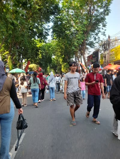 A street scene in Purwodadi, Indonesia, shows a crowd of people walking on a paved road lined with trees. The trees have full, green foliage and some have visible branches and trunks. On the right side of the road, there are buildings, some with yellow facades, and awnings or umbrellas. Some people are wearing casual clothing, including t-shirts, shorts, and jeans. Several individuals are wearing hijabs, indicating a likely Islamic cultural context. The lighting suggests it is daytime, possibly late afternoon given the angle of the sun and the shadows. Some individuals are looking at their phones. People are walking in both directions, suggesting a busy street or market area. There are also some stalls or vendors visible along the side of the road, with red and green umbrellas. The overall atmosphere appears to be that of a public gathering or a bustling street market.