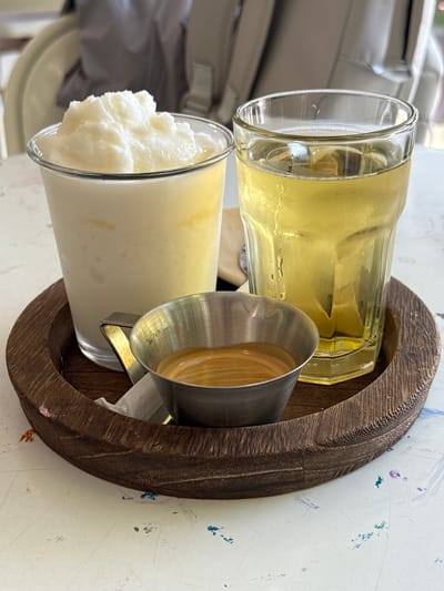 A close-up shot displays a wooden tray holding three items. On the left, a glass contains a creamy, white, frozen beverage with a textured surface. In the center, a small metal cup holds a dark, liquid substance, possibly coffee. To the right, a clear glass is filled with a light-yellow liquid, potentially iced tea. The tray and its contents are placed on a white surface with colorful paint splatters, suggesting an artistic environment. The background shows blurred figures and furniture, indicating an indoor setting. The overall impression is that of a cafe or restaurant in Tam Kỳ, Vietnam, offering a refreshing drink and a shot of espresso.