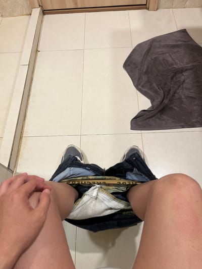 A first-person perspective shot shows a person sitting on a toilet in a tiled bathroom. The person is wearing jeans that are unzipped and partially pulled down, revealing underwear. Their hands are resting on their lap. To the right, a dark gray towel is draped on the floor. The bathroom features light-colored tile flooring and walls, with a wooden door frame visible in the background. The scene appears to be in a private residence.