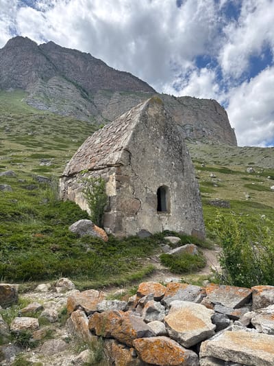 The image shows a stone structure with a small rectangular opening, resembling a mausoleum or a small tower, in a mountainous landscape. The structure is made of rough-hewn stones, with some areas appearing to be patched or repaired. Vegetation, including grass and some shrubs, grows around the structure and on the slopes of the surrounding mountains.

To the right of the structure, a pile of large, irregular stones forms a wall-like feature. These stones have an orangish-brown lichen growing on them.

In the background, a steep, rocky mountain face dominates the left side of the image. The mountain is striated with layers and appears to be made of dark gray rock. The sky above is partly cloudy, with patches of blue visible between white, puffy clouds. The lighting suggests it is daytime, likely midday or afternoon, with diffused sunlight.

The scene appears to be in a remote, natural setting, possibly in a historical or archaeological site given the architectural style of the stone structure. The overall mood is one of solitude and ancient history. There is no visible text in the image. The orientation of the image is rotated 90 degrees counter-clockwise from standard landscape orientation.