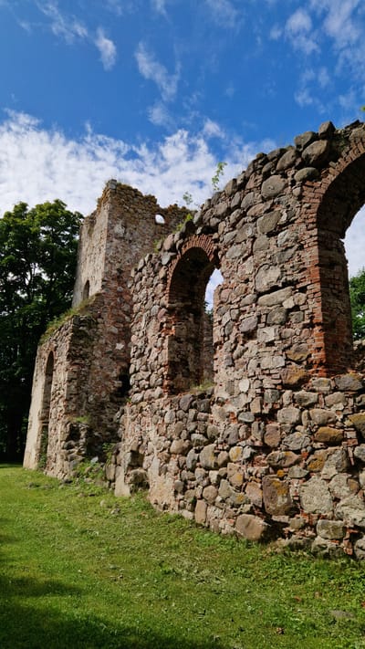 A low-angle, full shot captures the weathered stone ruins of the Embūte Evangelical Lutheran Church against a vibrant blue sky dotted with scattered white clouds. The ancient structure is composed of large, irregularly shaped stones and red brick, with arched openings visible, hinting at its former grandeur. Green grass covers the ground in the foreground, with a large, dark green tree on the left side of the frame. The scene is bathed in bright sunlight, casting shadows on the grass and highlighting the textures of the stonework.