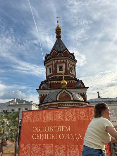 The image shows a red brick church with gold domes against a blue sky with white clouds and a contrail. In the foreground, an orange banner with decorative patterns and Russian text, "ОБНОВЛЯЕМ СЕРДЦЕ ГОРОДА" (Updating the Heart of the City), is visible. A woman with blonde hair in a light yellow t-shirt and blue jeans is partially visible on the right side of the frame, looking away from the camera. Buildings with grey roofs are seen behind the banner and to the left of the church. The scene appears to be an outdoor public space.