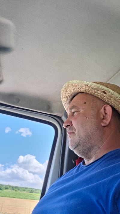 In Mereșeni, Moldova, a middle-aged man wearing a straw hat and a blue t-shirt is seen from the right side, looking out of a vehicle window. The interior of the vehicle is visible at the top of the frame, with a gray ceiling. Outside the window, a bright blue sky with scattered white clouds is visible, along with a distant green landscape. The man has a mole on his cheek and a lightly stubbled chin. The lighting suggests it is daytime.