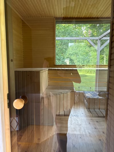 The image shows the interior of a wooden sauna with a large window. The sauna has wooden benches and a wooden floor. Through the window, a green, grassy area with trees can be seen. A wooden door handle is visible on the left side of the image. The overall scene appears to be calm and serene. The location is a private residence in Jaunbagumi, Latvia.