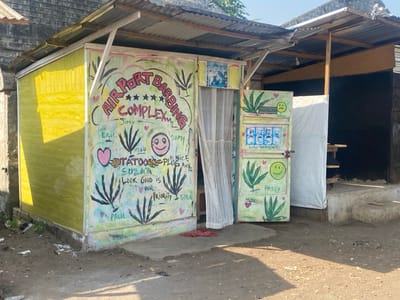 This media file captures an informal, brightly painted barbershop structure in what appears to be a local commercial area, likely in Victoria, Cameroon. No people are visible in the scene.
The primary subject is a small, makeshift building featuring a yellow side wall and a white facade, both constructed from simple materials. A corrugated metal roof provides shelter for this structure and an adjacent, darker building to the right. The ground is unpaved, consisting of dirt and gravel with some scattered debris.
The white facade of the barbershop is covered in colorful, hand-painted graffiti-style artwork and text. Dominating the upper section are the words "AIRPORT BARBING COMPLEX" arched over several stars. Below this, various names are inscribed, including "Jimy," "ERIC," "SAMY," "HERU," "SUZAA," "PAUL," and "BOBA." Other phrases visible are "TATOO PLUG NICE PAYZ" and "LOOK GOOD IS OUR PRIORITY." The wall also features a pink heart, a smiley face, and several stylized leaf designs. A light-colored, patterned curtain serves as the entrance to the barbershop.
To the right, a freestanding, detached door (possibly repurposed from a refrigerator) is also vibrantly painted. It displays the name "HUNCHI" next to a sad face, and "PASSY" alongside a smiley face. The door also features multiple images of faces labeled "LIL WAYNE" and is decorated with prominent cannabis leaf motifs.
The scene is illuminated by bright daylight, casting distinct shadows on the ground, indicating either morning or late afternoon sun under clear skies. The overall impression is one of a lively, community-oriented business, characterized by its unique, artistic expression and informal construction.