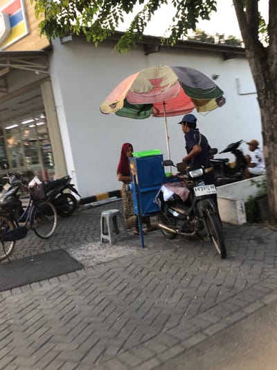 In Sentul, Indonesia, a man wearing a dark blue shirt and a blue baseball cap stands behind a mobile food cart attached to a motorcycle. The cart has a blue metal body and a faded, multi-colored umbrella providing shade. A woman wearing a red headscarf and a patterned skirt stands to the left of the cart. A small grey stool is positioned in front of the cart. In the background, several motorcycles are parked, and another person wearing a hat and a white shirt is seated. To the left of the scene, a blue bicycle is parked. The environment appears to be a street or sidewalk with a brick-paved ground. A tree with a thick trunk is visible on the right side of the image. The scene is likely outside a commercial establishment, suggested by the building and signage partially visible on the left.