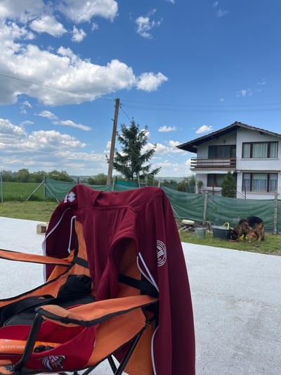 The image depicts an outdoor scene in Klenovik, Bulgaria on a sunny day with a blue sky and scattered clouds. In the foreground, a red tracksuit jacket and orange bag hang on a black structure of the type used to support a table or chair. The jacket features a white logo near the left chest area. Behind this, a two-story white house is visible, partially obscured by a green fence. A large German Shepherd dog is visible in the yard to the right of the house. A wooden utility pole stands nearby, with power lines stretching across the frame. Beyond the fence, there is a grassy field and trees.