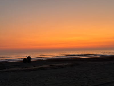A tranquil beach scene unfolds at sunset. The sky is painted with gradients of orange and pink hues. The ocean meets the shore with gentle waves. Silhouetted against the bright horizon, a group rides a vehicle on the dark sand. Tire tracks mark the beach surface. The scene evokes a sense of serenity and peaceful activity.