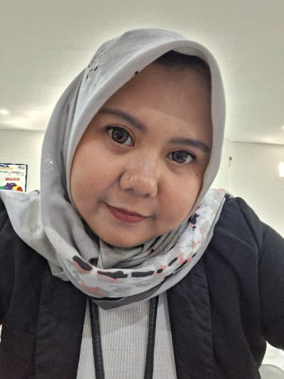 A person wearing a light gray hijab with subtle pink and black patterns looks directly at the camera. She is also wearing a black jacket over a white striped shirt. The background is a blurry white wall with what appears to be a colorful poster near the bottom of the frame. The lighting is bright and even.